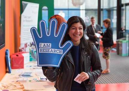 A female researcher holding up a blue foam hand that says 'I am research'